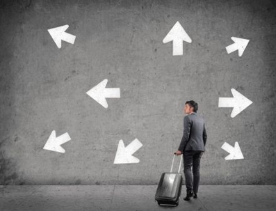 An uncertain man with a rolling suitcase stands in front of a blank wall with white arrows all around him pointing in different directions.